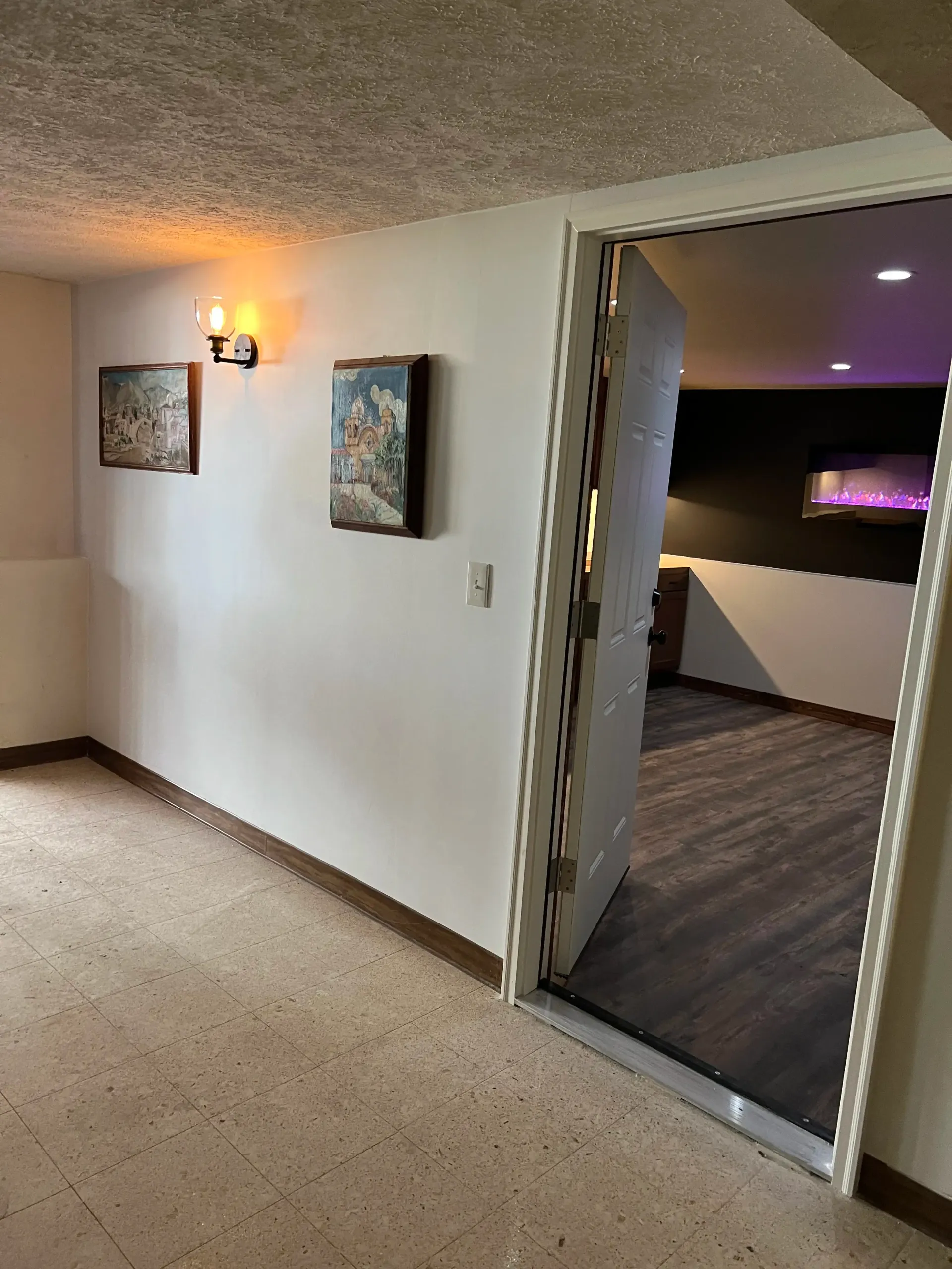 A hallway with speckled flooring, two framed pictures on a wall with a lit sconce, and an open doorway to another room.