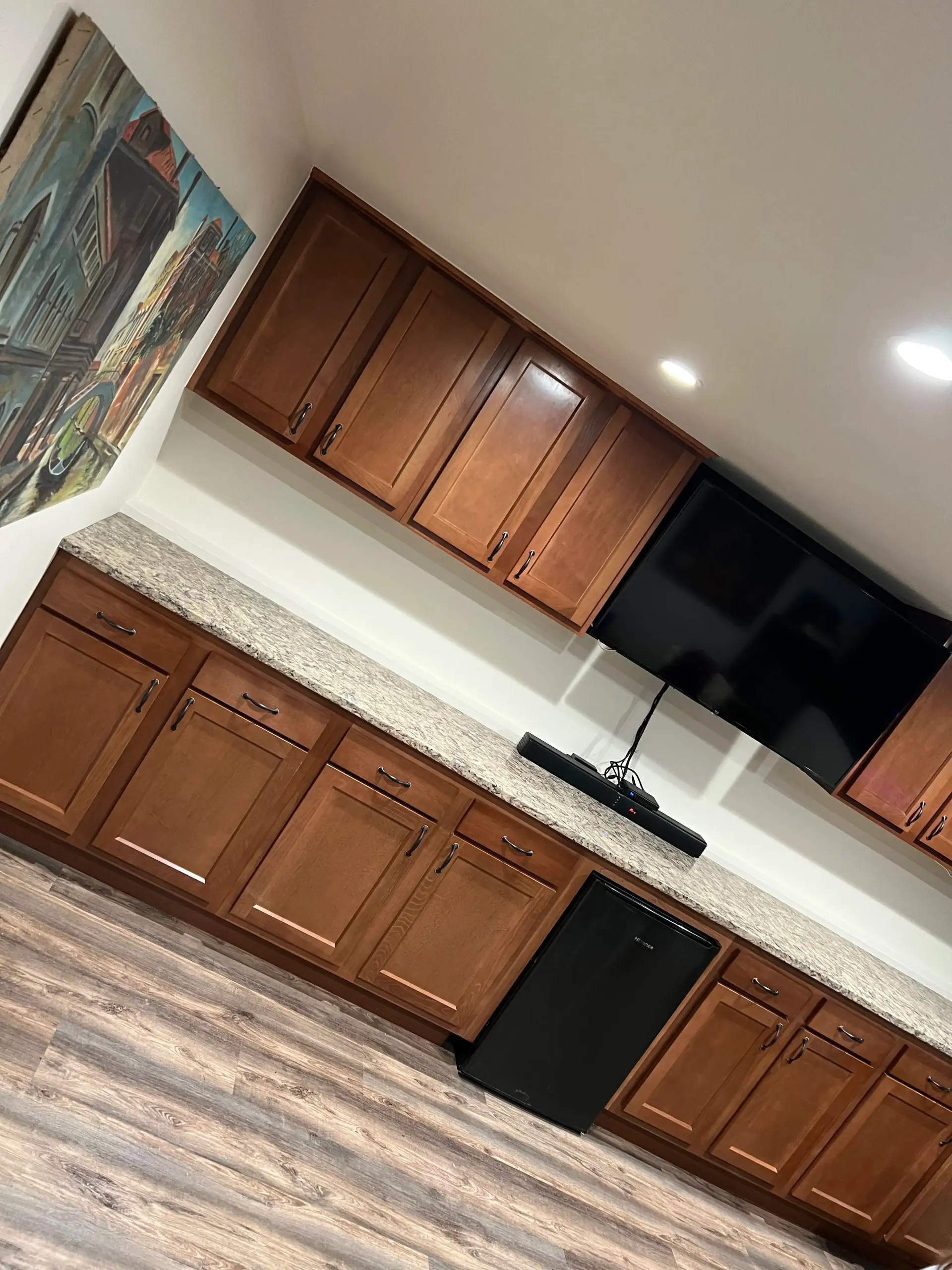 A home bar with wooden cabinets, speckled granite countertops, a built-in black mini fridge, and a wall-mounted TV.