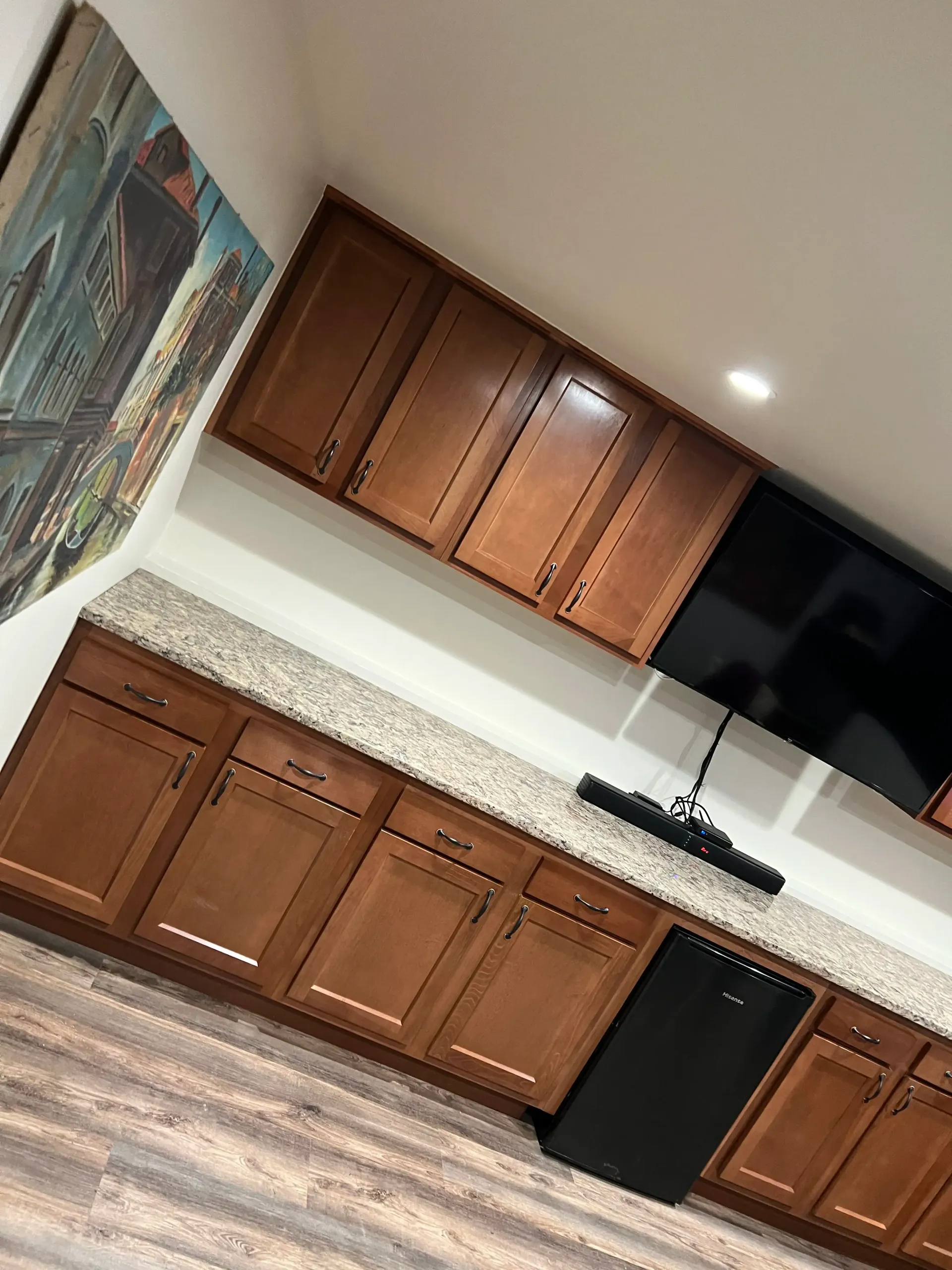 A room featuring wooden cabinets, a granite countertop, a wall-mounted TV with a soundbar, and wood-look flooring.