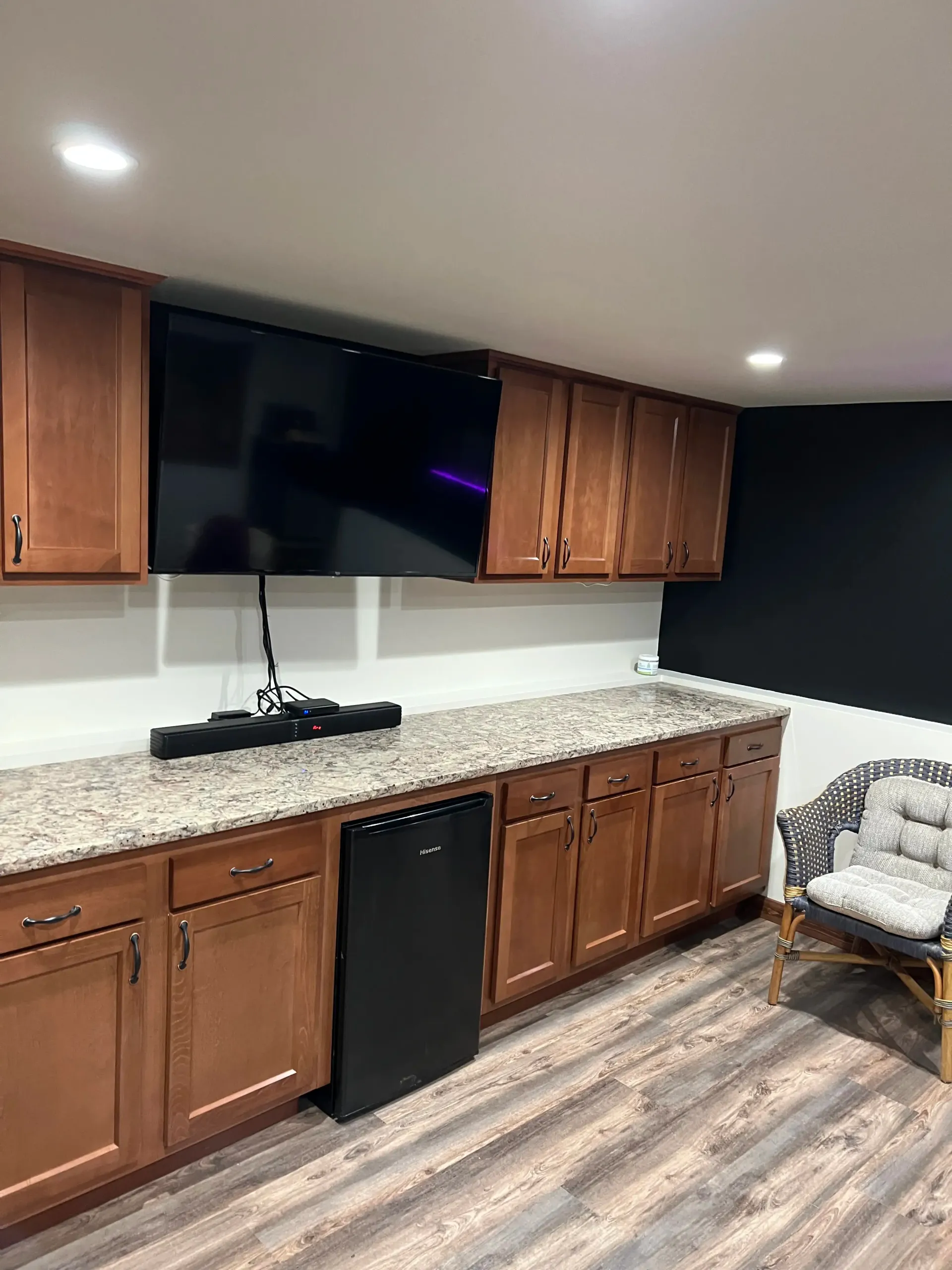 A kitchenette with wood cabinets, granite countertops, a black mini fridge, a mounted TV, and a wicker chair.