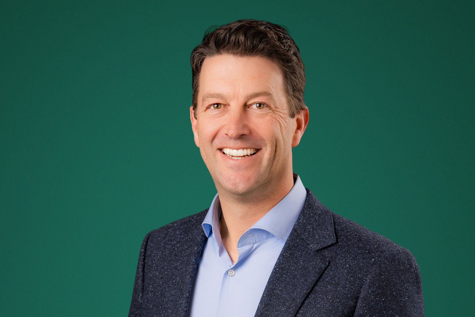 Man in blue button-down shirt and patterned jacket smiling against a green background.