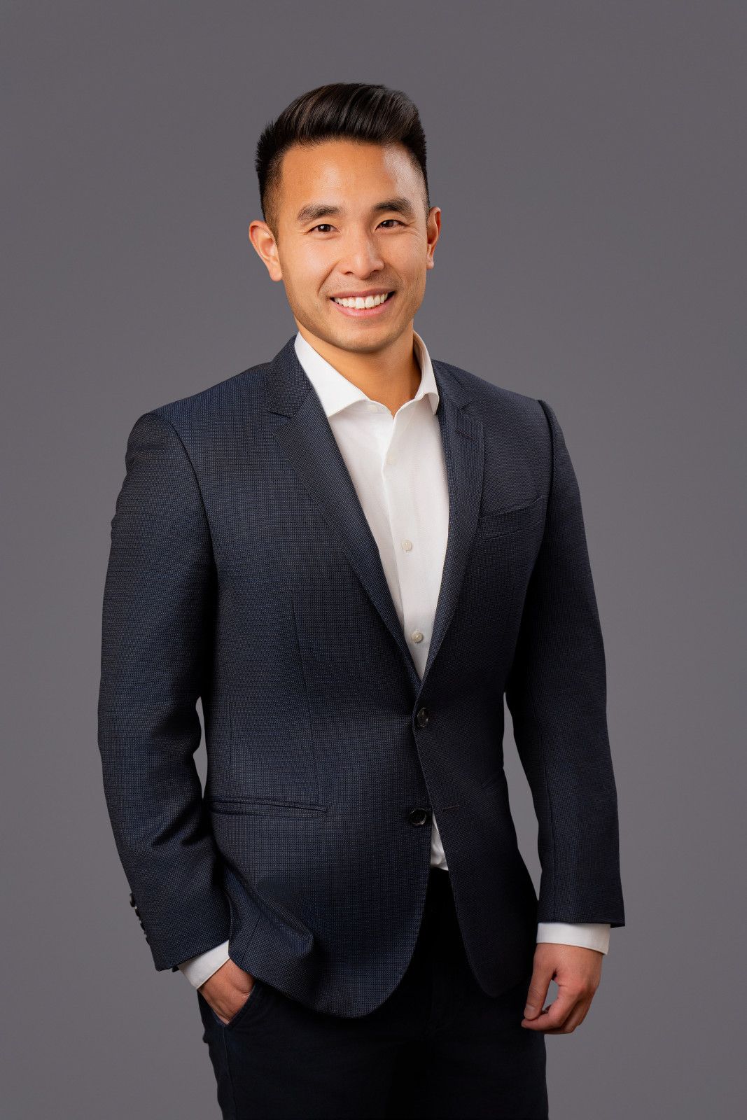 Man in dark blue blazer and white shirt smiles, hands in pockets against gray backdrop in Newport Beach office