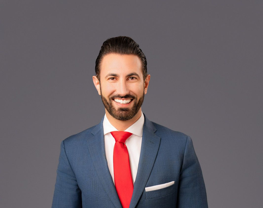 A man with a beard, wearing a blue blazer, white shirt, and red tie, smiling against a plain gray background. 