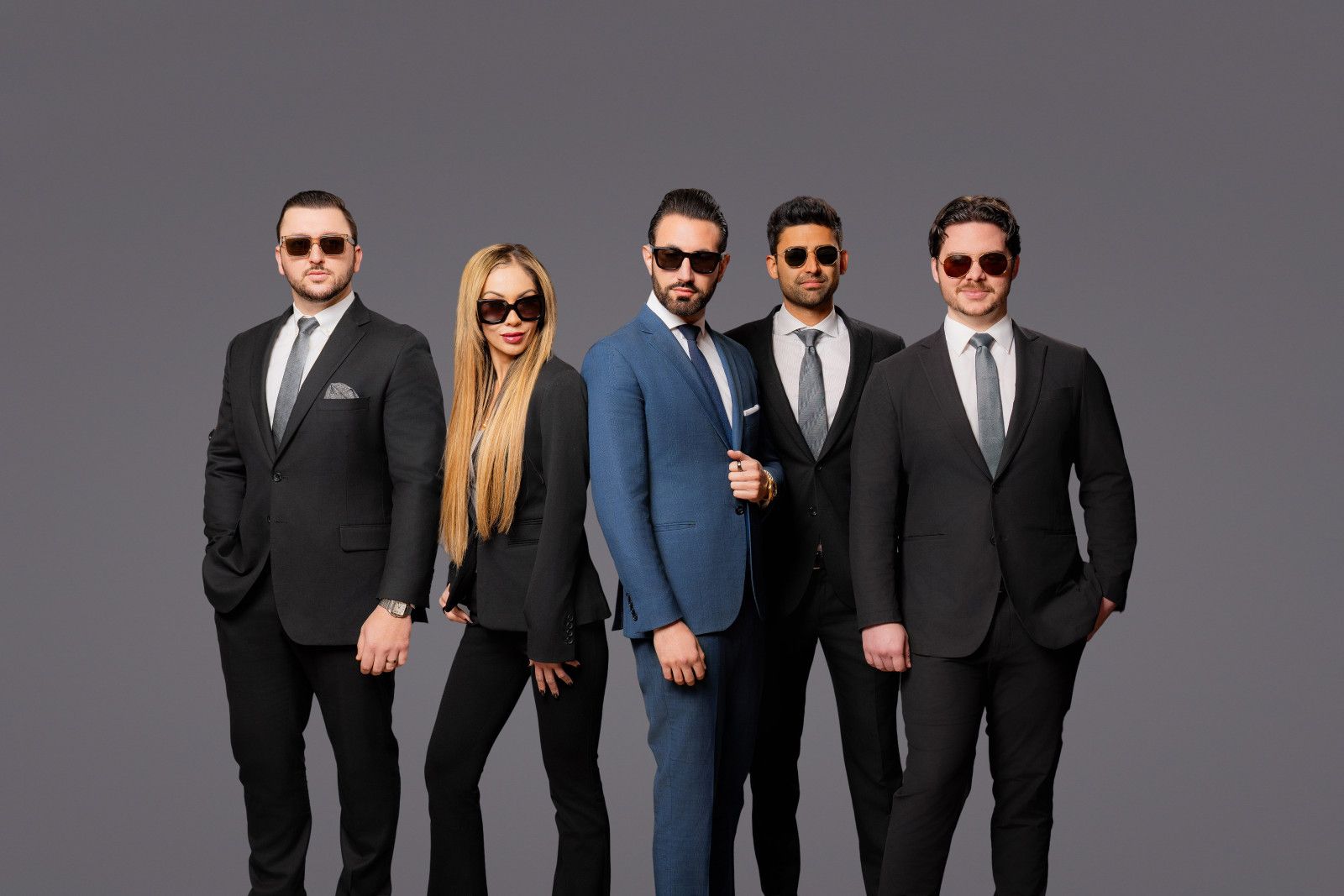 Five people in professional business suits and sunglasses standing in a line against a gray background.