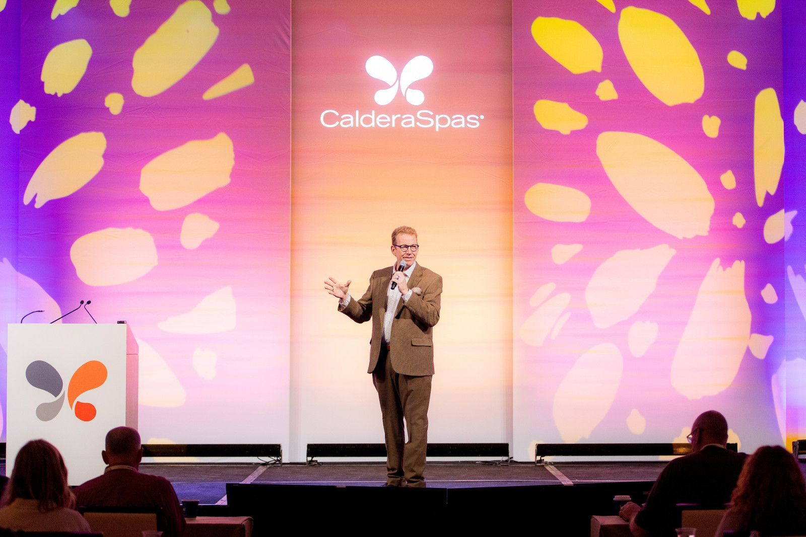 A person in a suit speaks on a stage with a Caldera Spas logo and purple, orange, and yellow floral-patterned backdrops.
