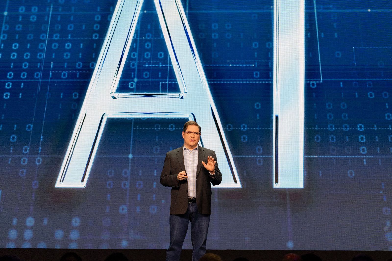 Orange County conference photo A person stands on a stage in a dark blazer before a large, blue, glowing screen displaying the letters