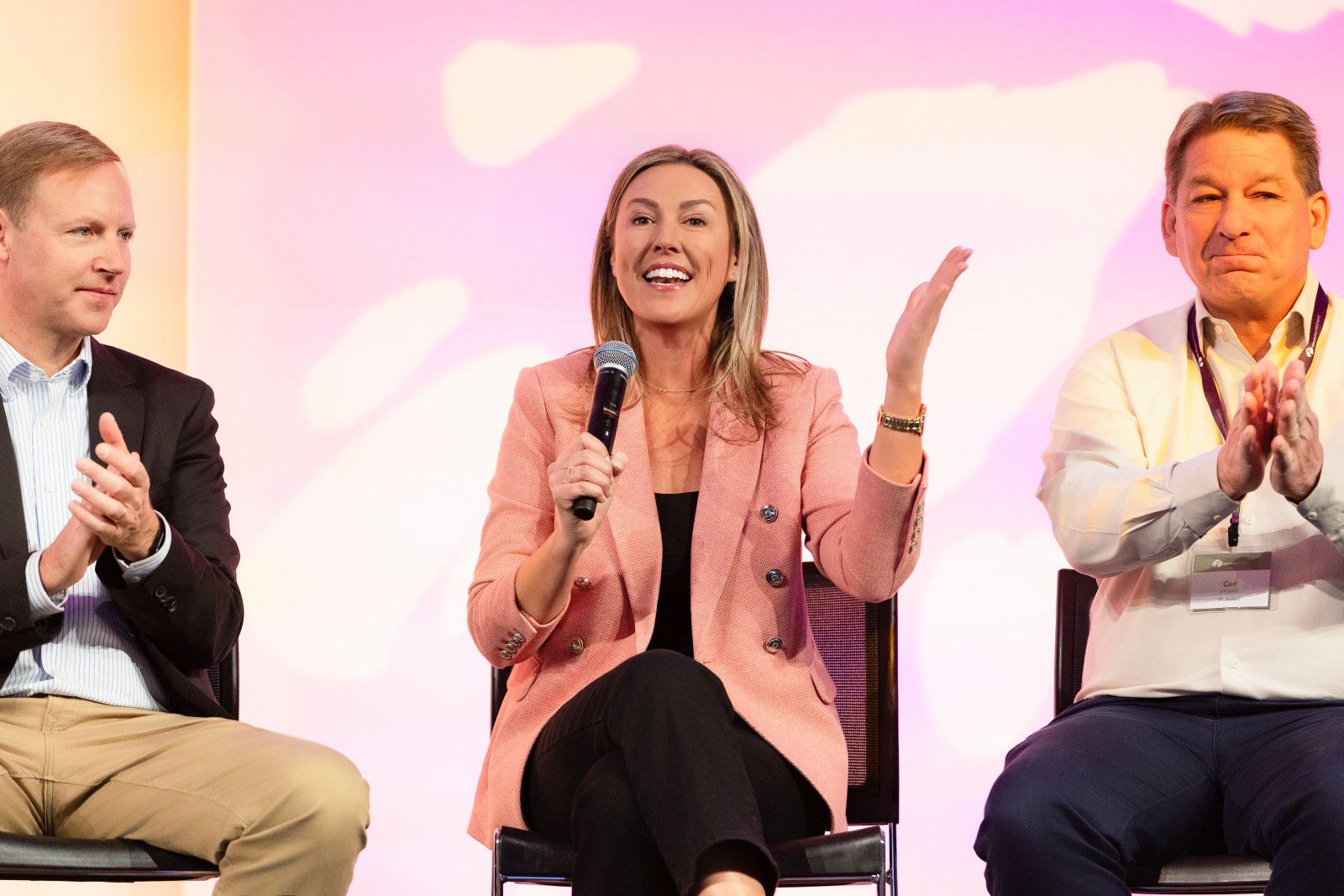 orange county conference photographer A woman in a pink blazer speaks into a microphone while two men sit on either side of her, applauding on a stage at a conference in Orange County
