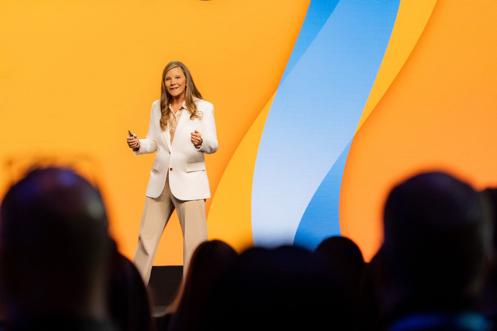 A speaker stands on stage in front of a bright orange and blue background, gesturing while addressing an audience.