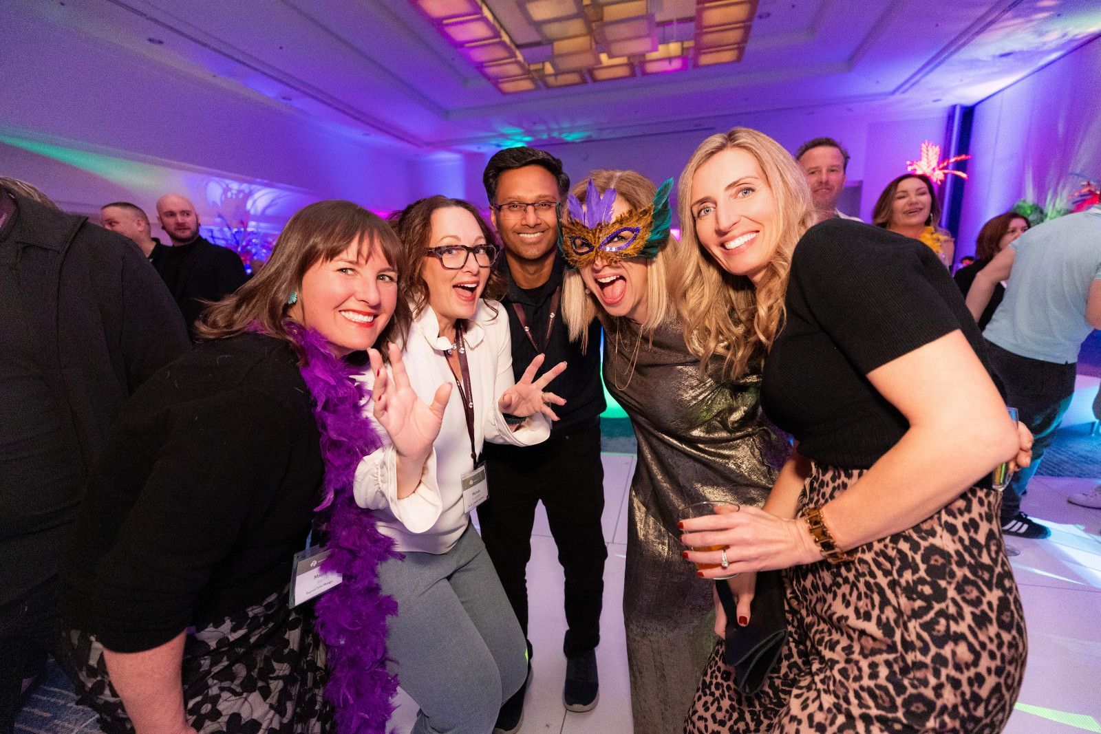 conference-convention-photographer-orange-county A group of friends laughing and posing at an indoor evening event, some wearing festive masks and purple feather boas.