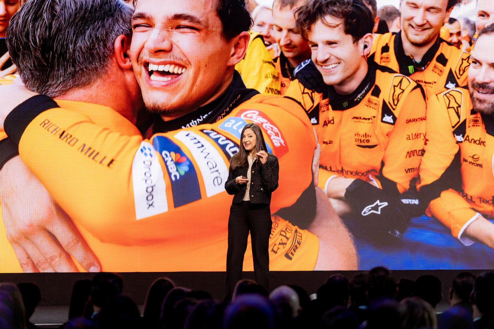 key-note speaker at conference in Anaheim A person stands on a stage in front of a large screen showing a Formula 1 team celebrating in orange racing suits at a convention