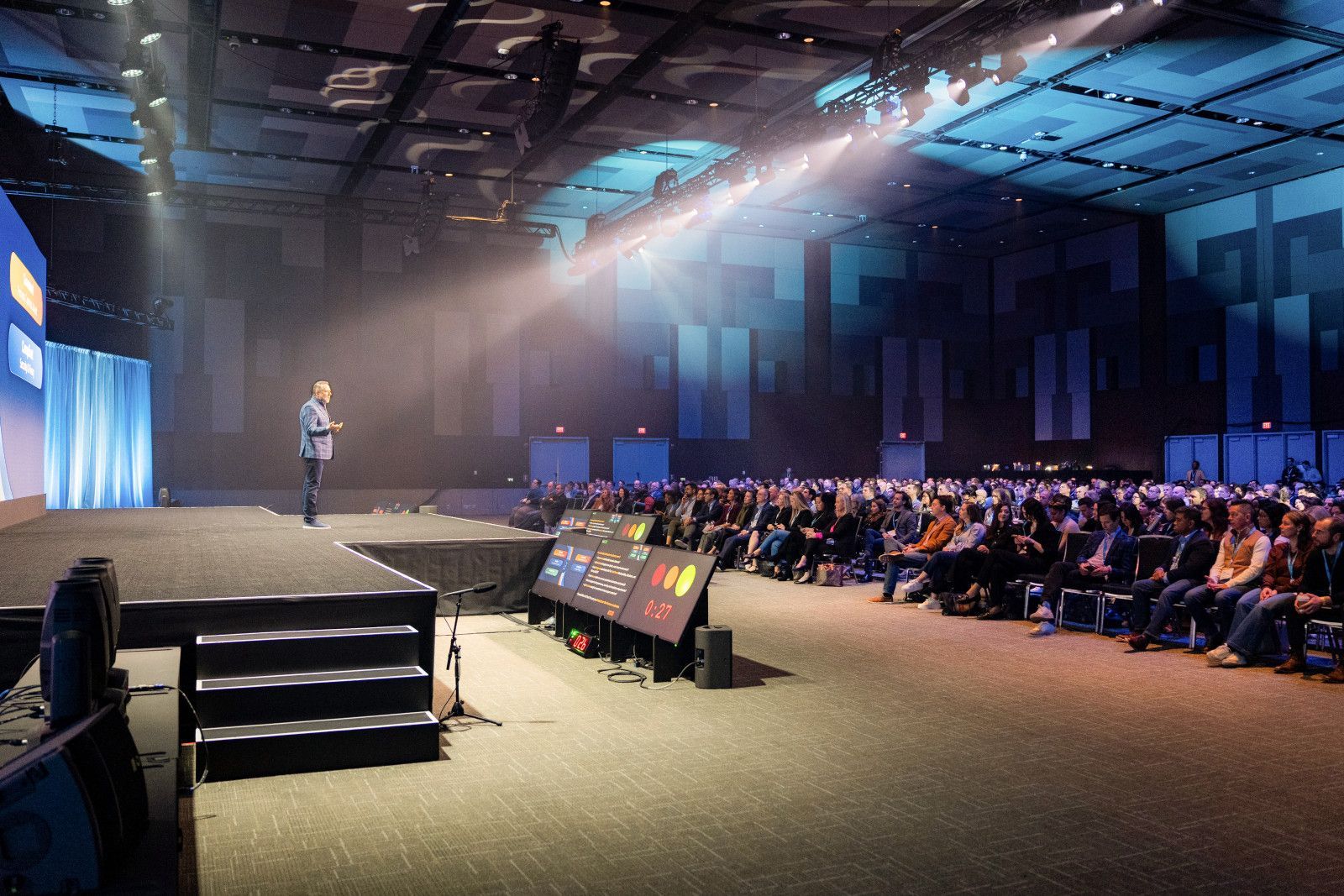 Anaheim Convention Center photographer A speaker stands on a stage bathed in spotlight, presenting to a large audience seated in a dark, expansive conference hall at Anaheim Convention Center