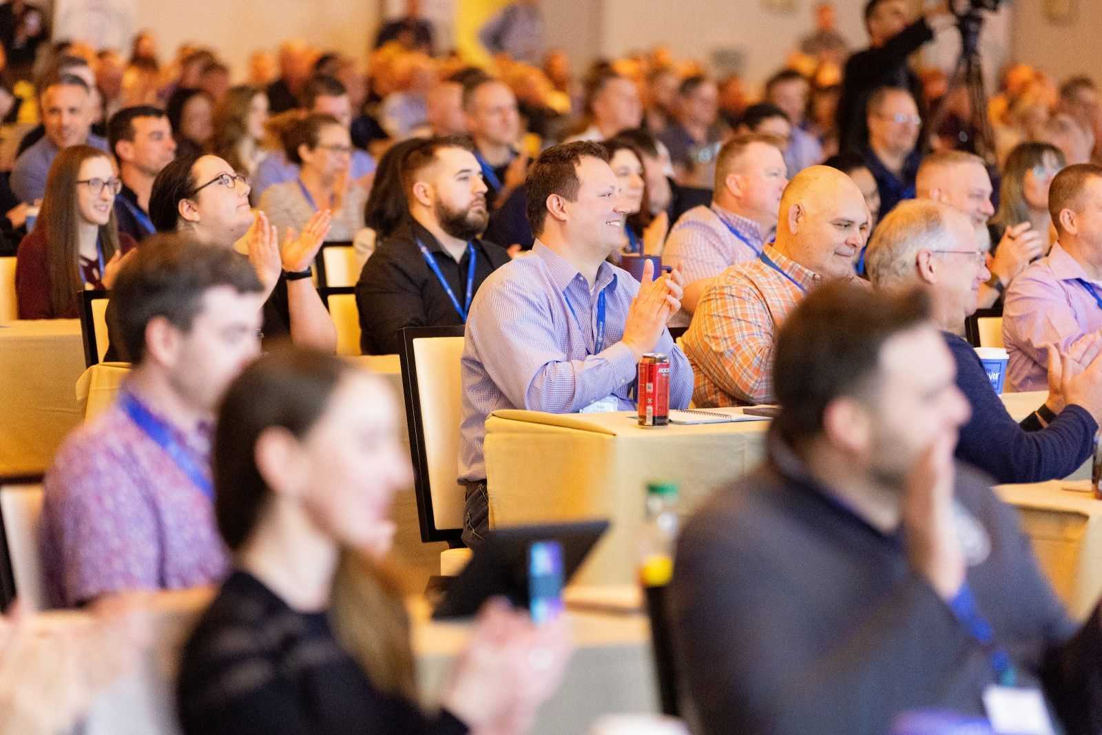 A large, diverse group of people sitting at tables in a conference hall, listening to a presentation and clapping.
