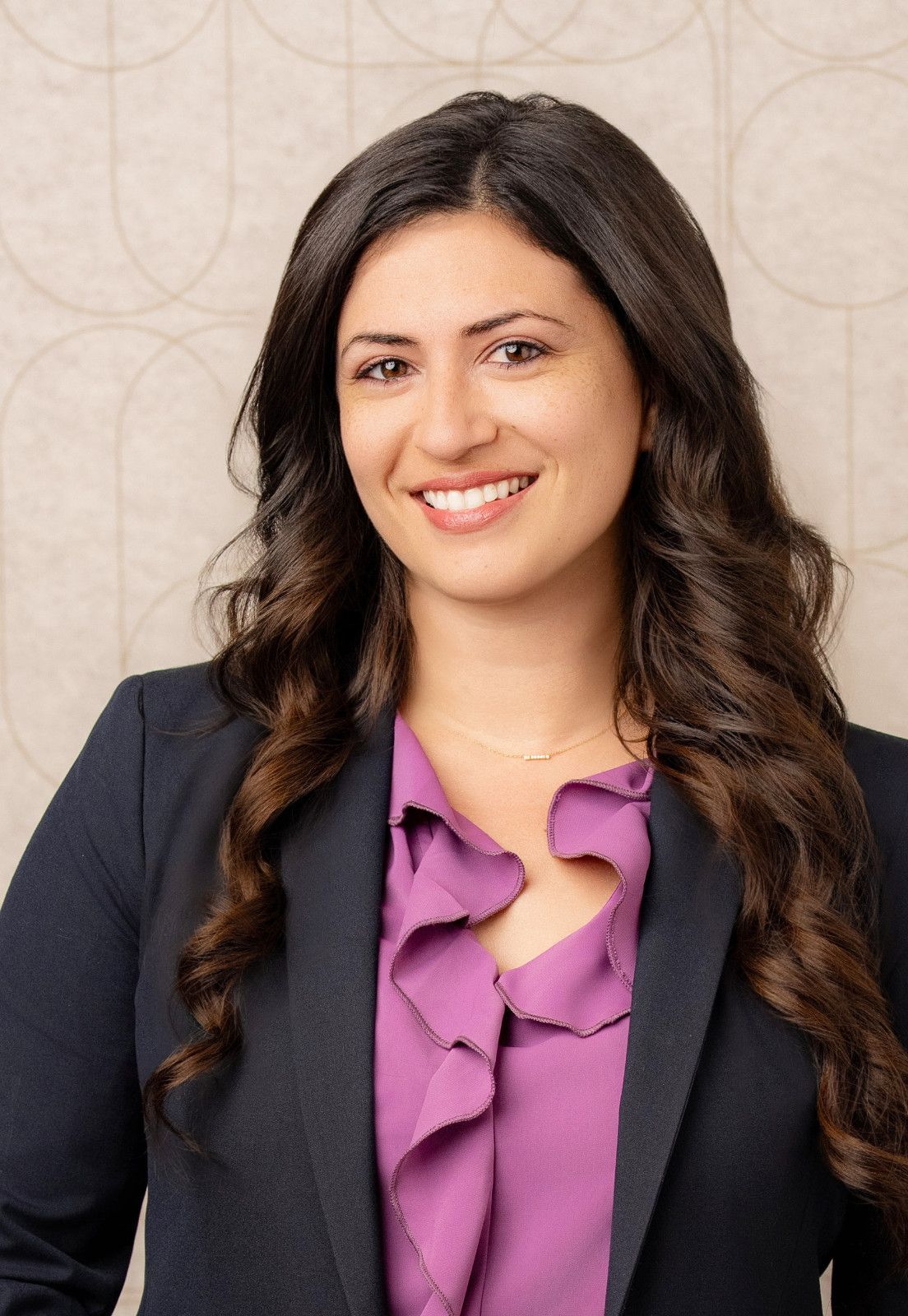 female corporate headshot on-site photographer A professional headshot of a woman smiling, wearing a dark blue blazer over a purple ruffled blouse, against a light wall in orange county