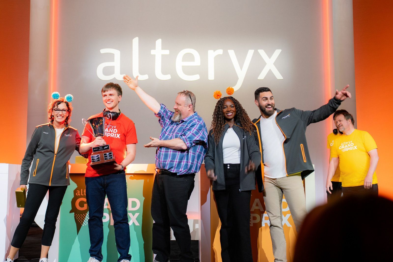 People on stage in front of an Alteryx logo, with one person holding a trophy and others gesturing toward the audience.