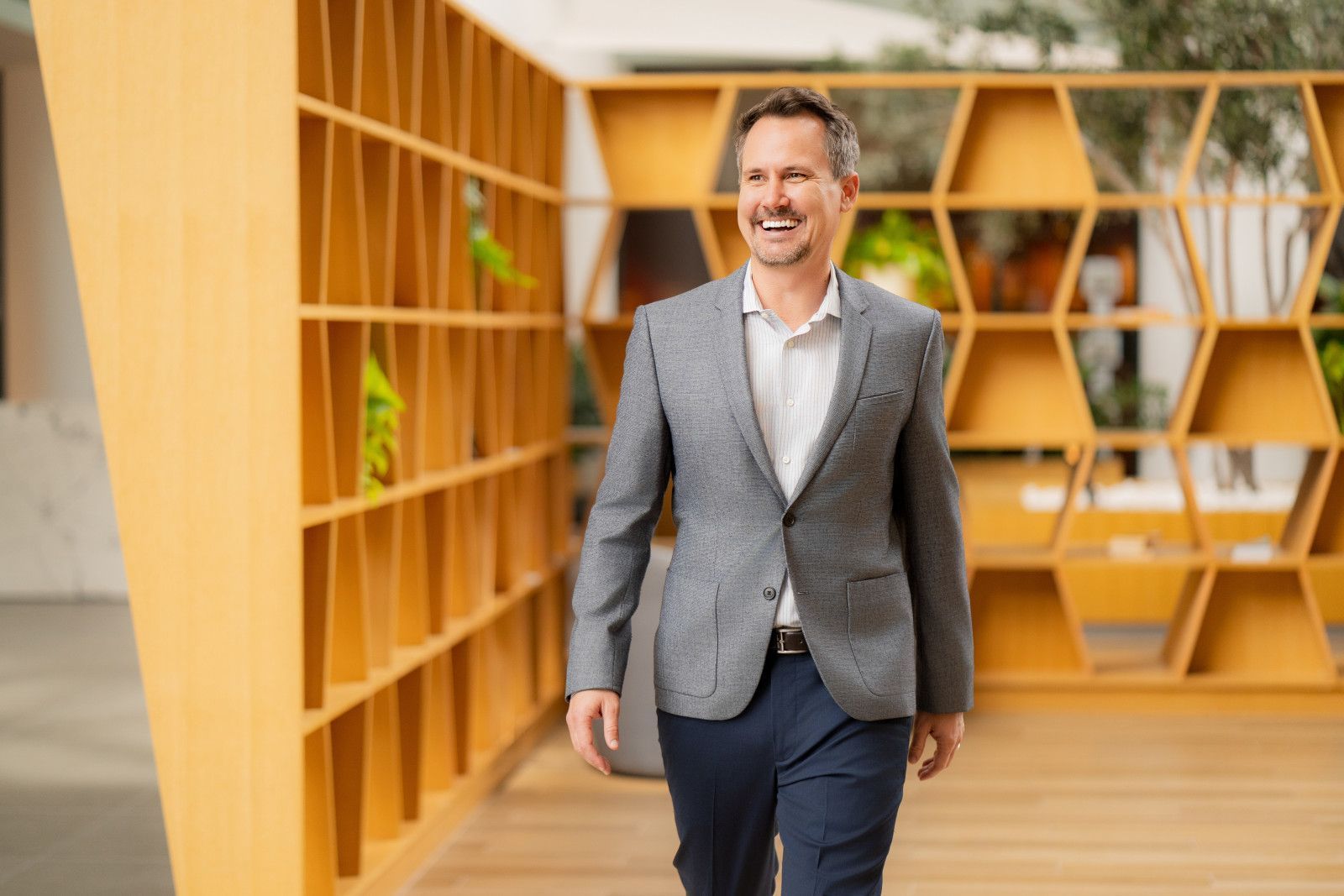 Man in a gray blazer smiling, walking towards the camera in a modern office.