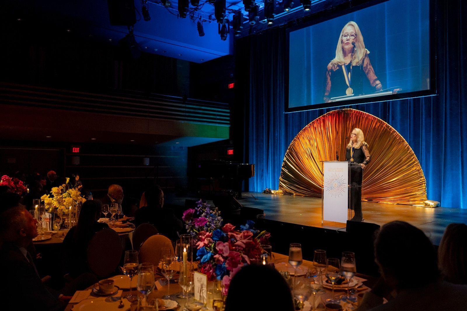 A speaker stands at a podium on a stage with a gold backdrop, with their image projected on a large screen above.