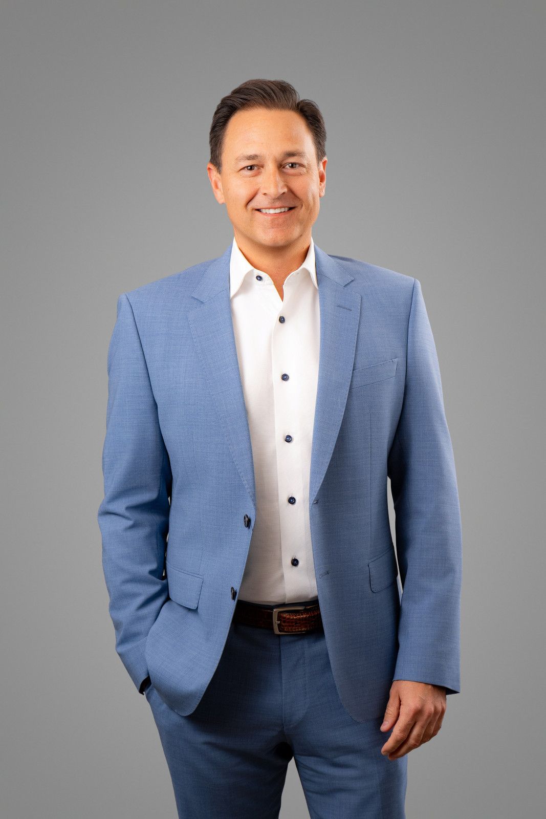 Corporate Headshot Portrait Anaheim Man in blue suit, white shirt, smiling, hands in pockets, gray background in Anaheim Office