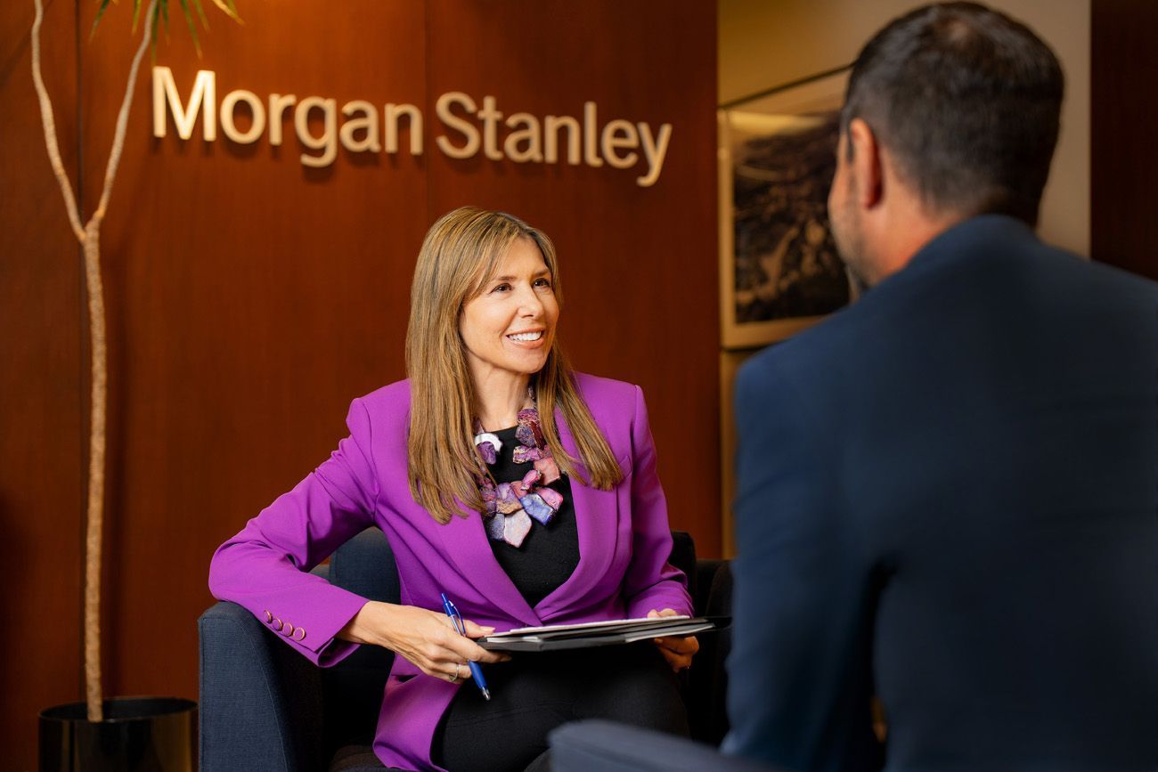 Woman in purple blazer, holding a tablet, talking to a man in a Morgan Stanley office.
