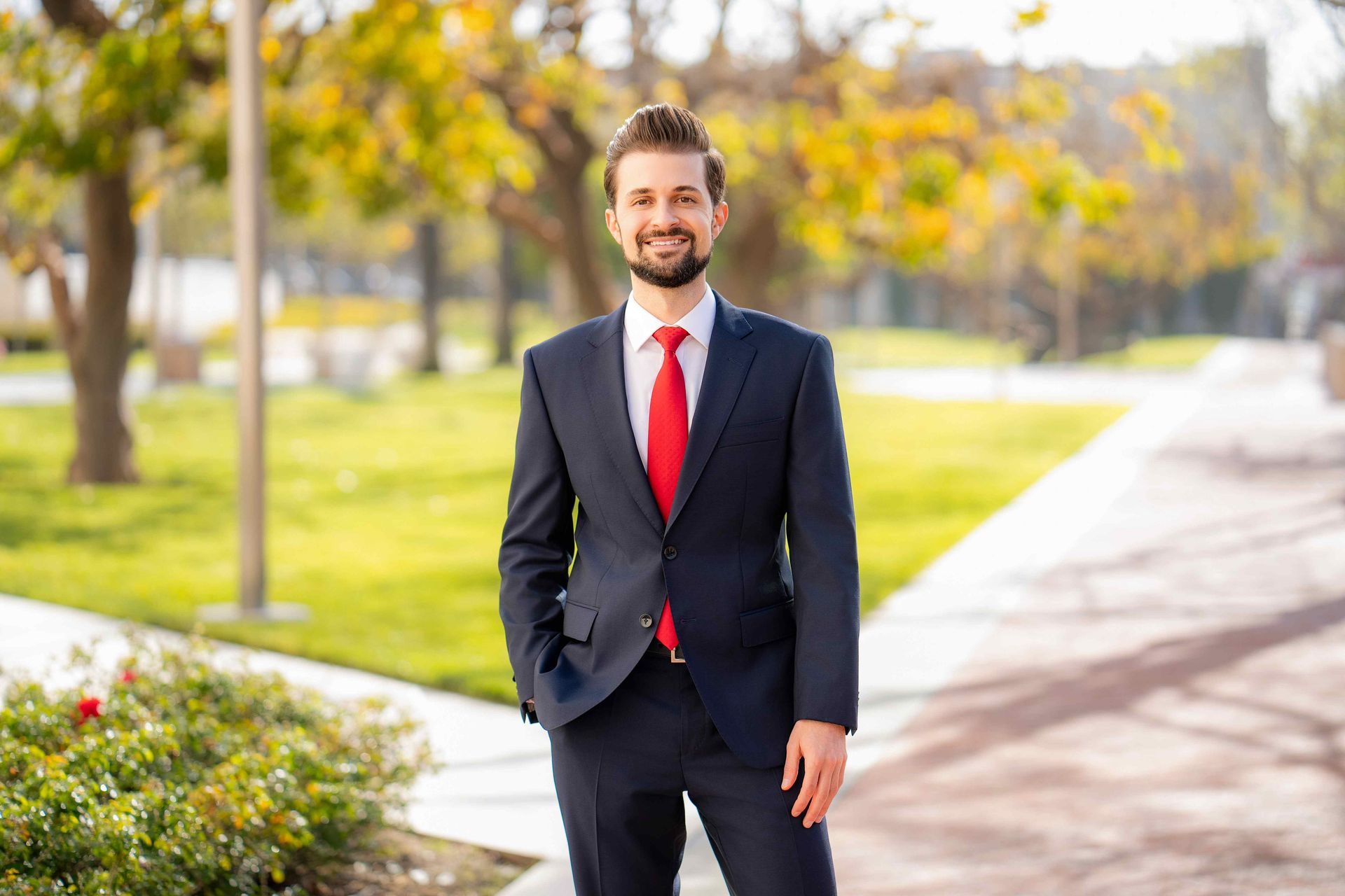 A man in a suit and red tie is standing in a park.