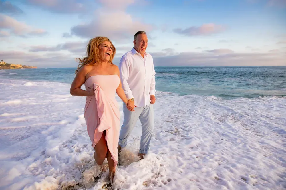 A man and a woman celebrate getting engaged walking on the beach holding hands.