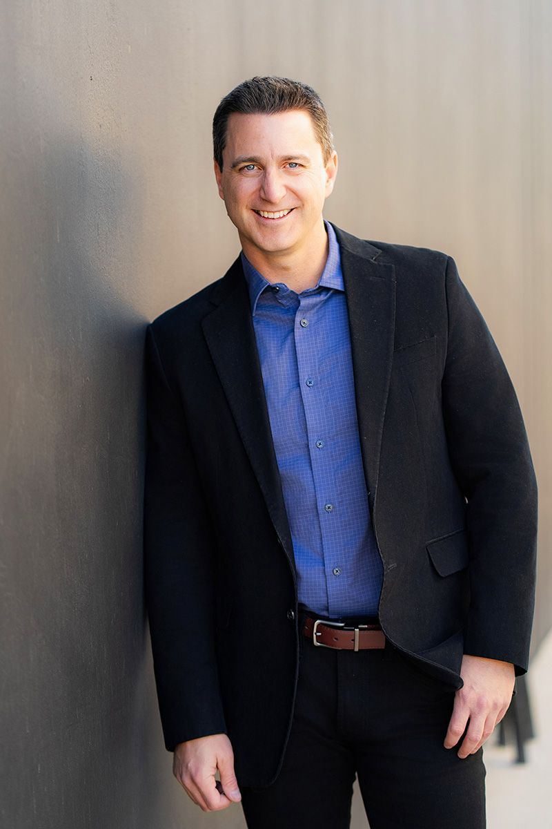 A man in a suit and blue shirt is standing in front of a wall for a Hinge App photo