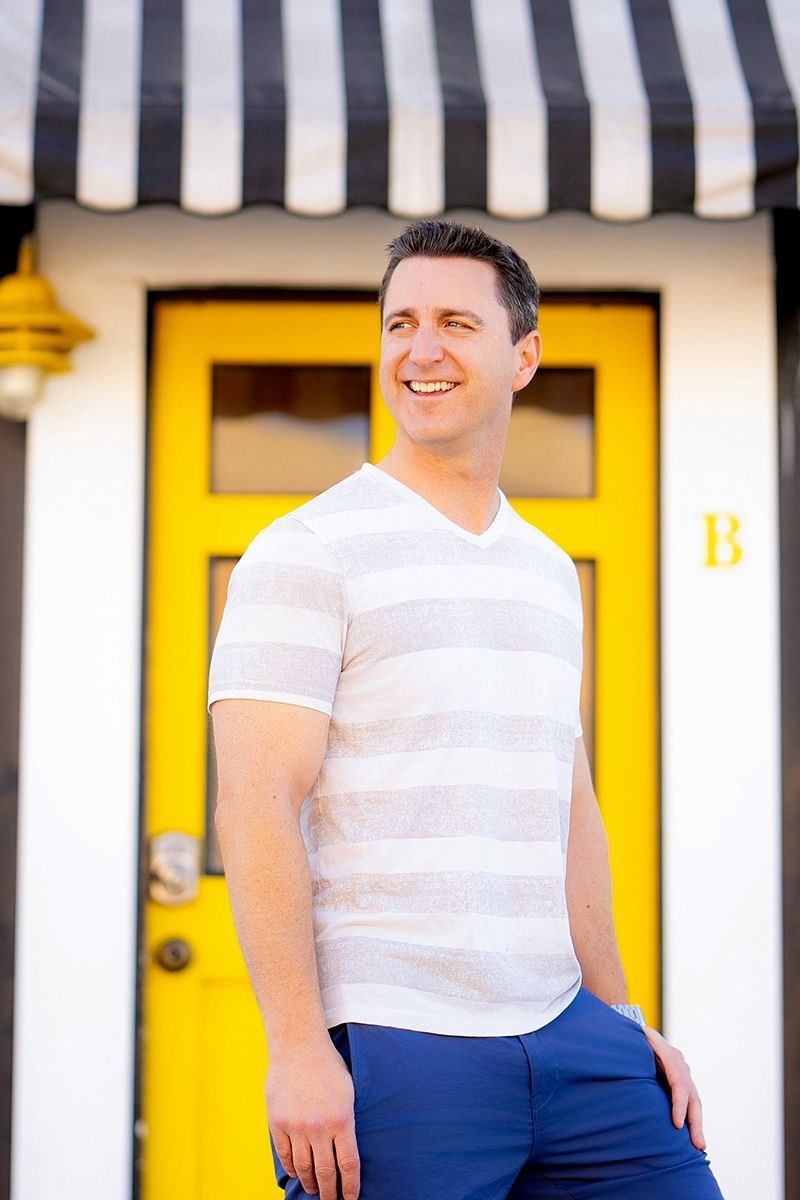A man in a striped shirt is standing in front of a yellow door for a Bumble App photo.