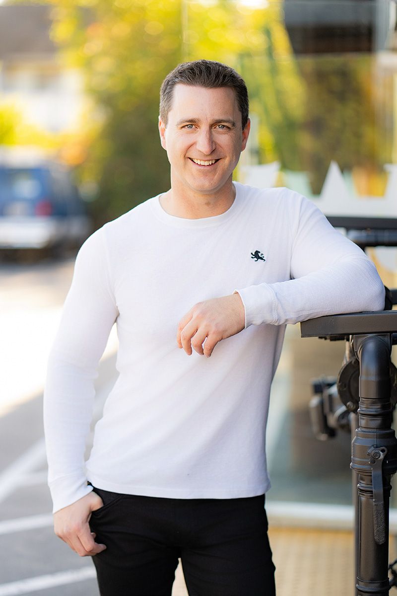 A man in a white sweater and black pants is leaning against a railing for a Tinder photo