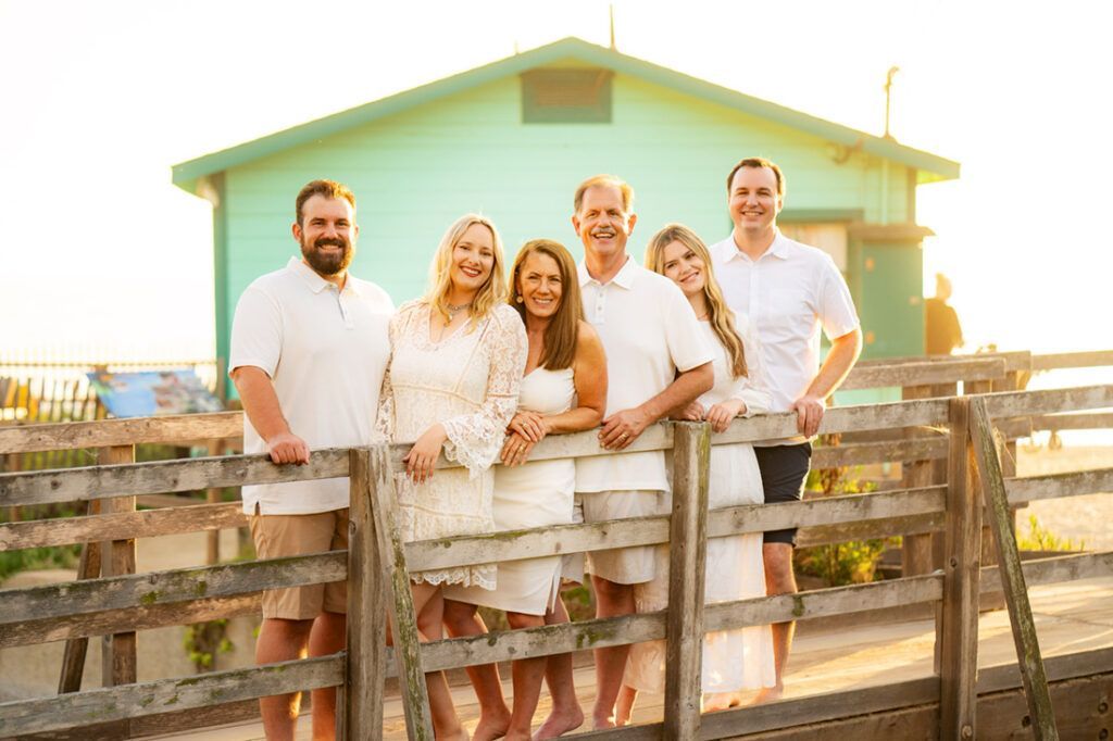 A family is standing on a wooden bridge in front of a green beach cottage.