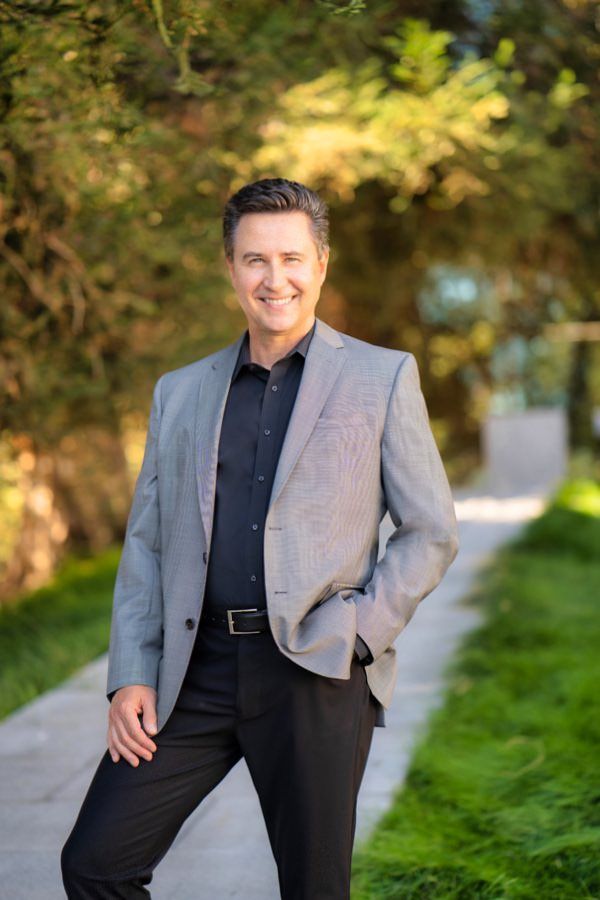A man in a suit poses for a headshot in front of a park