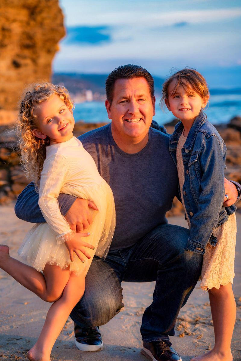 Laguna Beach Family Photo A dad is kneeling with his young daughters on a beach.