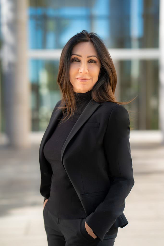A woman in a black jacket and turtleneck is standing in front of a building with her hands in her pockets.