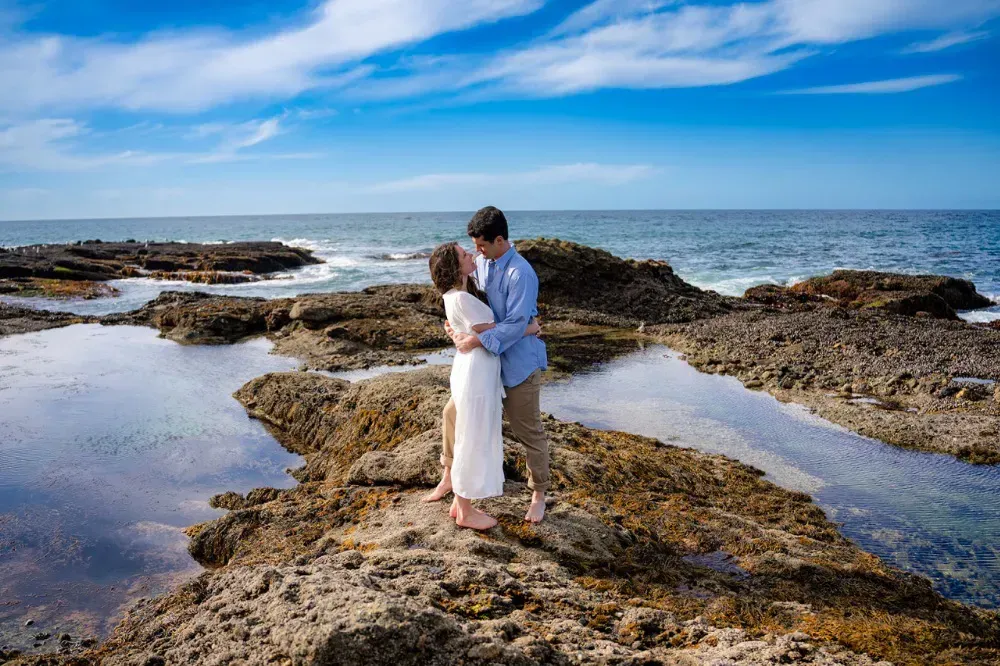 A man and woman are kissing on a rocky beach near the ocean.