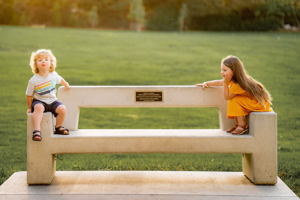A boy and a girl are sitting on a bench in a park.