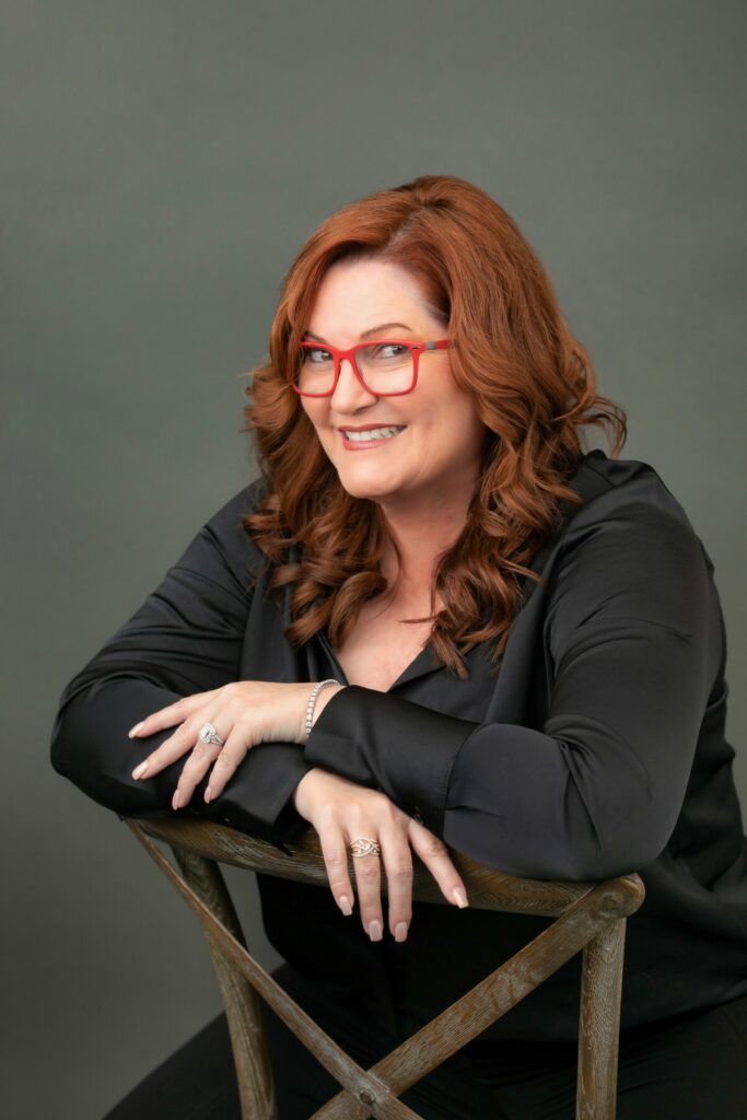 A woman with red hair and glasses is sitting on a chair for a LinkedIn photo