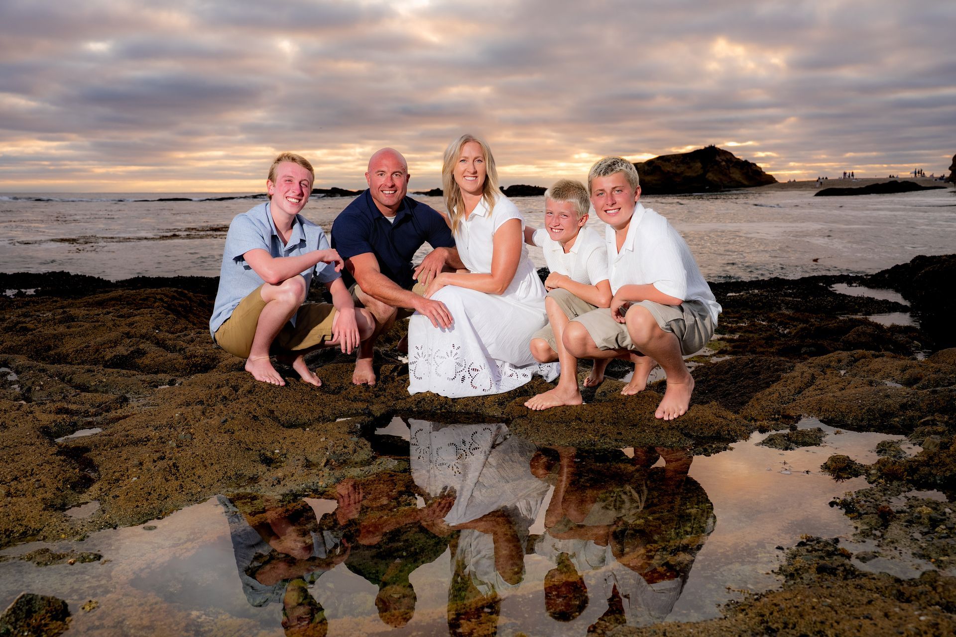 Treasure Island Beach Family Portrait Family of five crouches on rocks, ocean backdrop. Sky with orange hues reflects in water at Treasure island beach in laguna