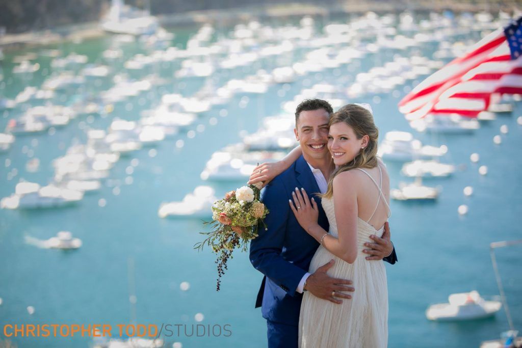 A bride and groom are posing for a picture in front of Avalon Harbor in Catalina