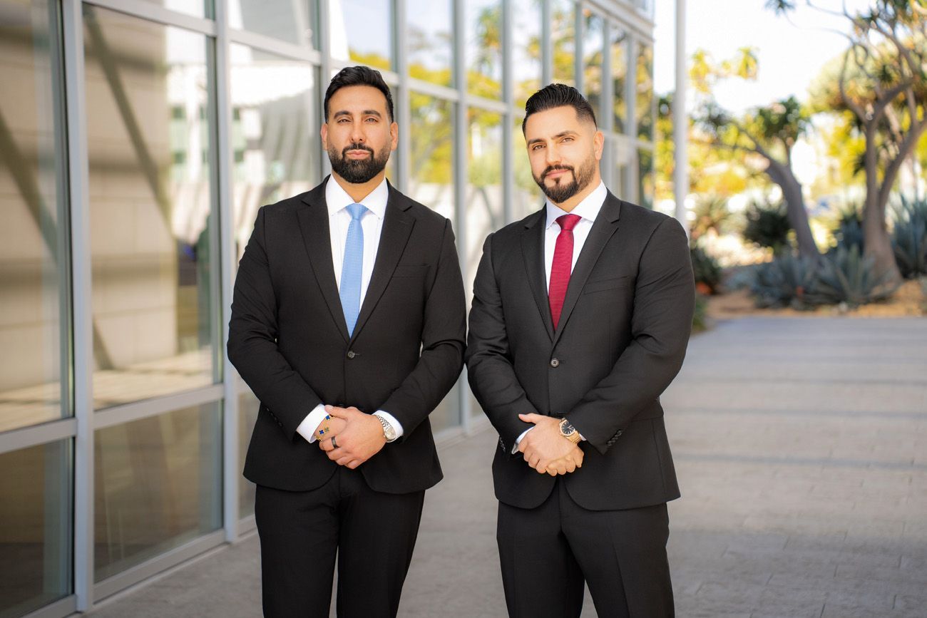 attorney brand photo in Irvine Two men in dark suits and dress shirts stand side-by-side outside a glass building, hands clasped, looking at the camera.