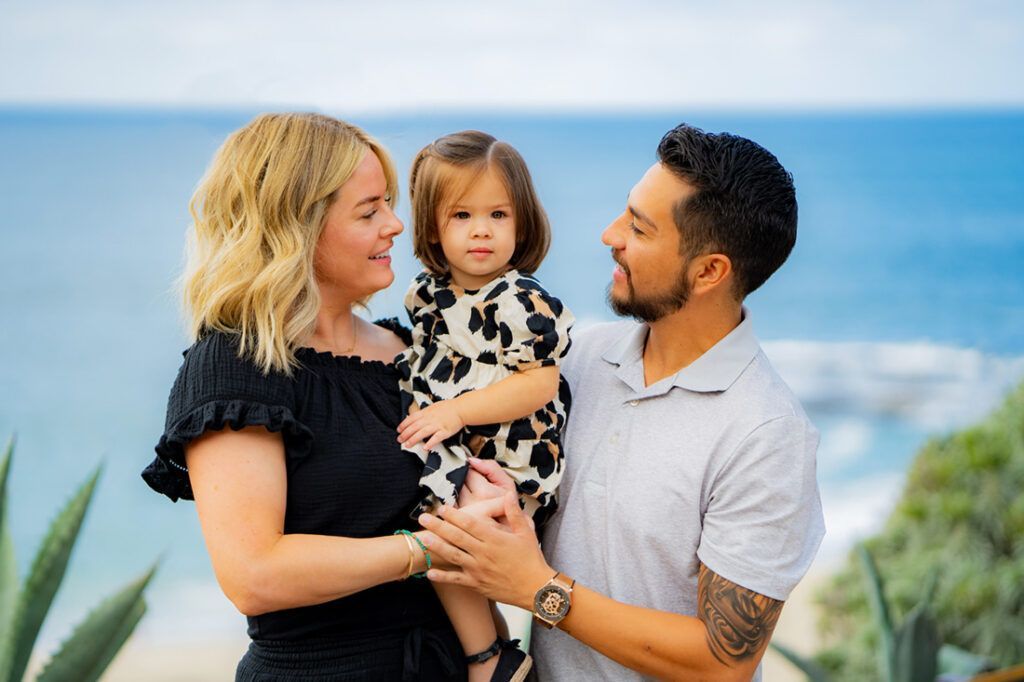 A man and woman are holding a little girl in front of the ocean In Newport Beach.