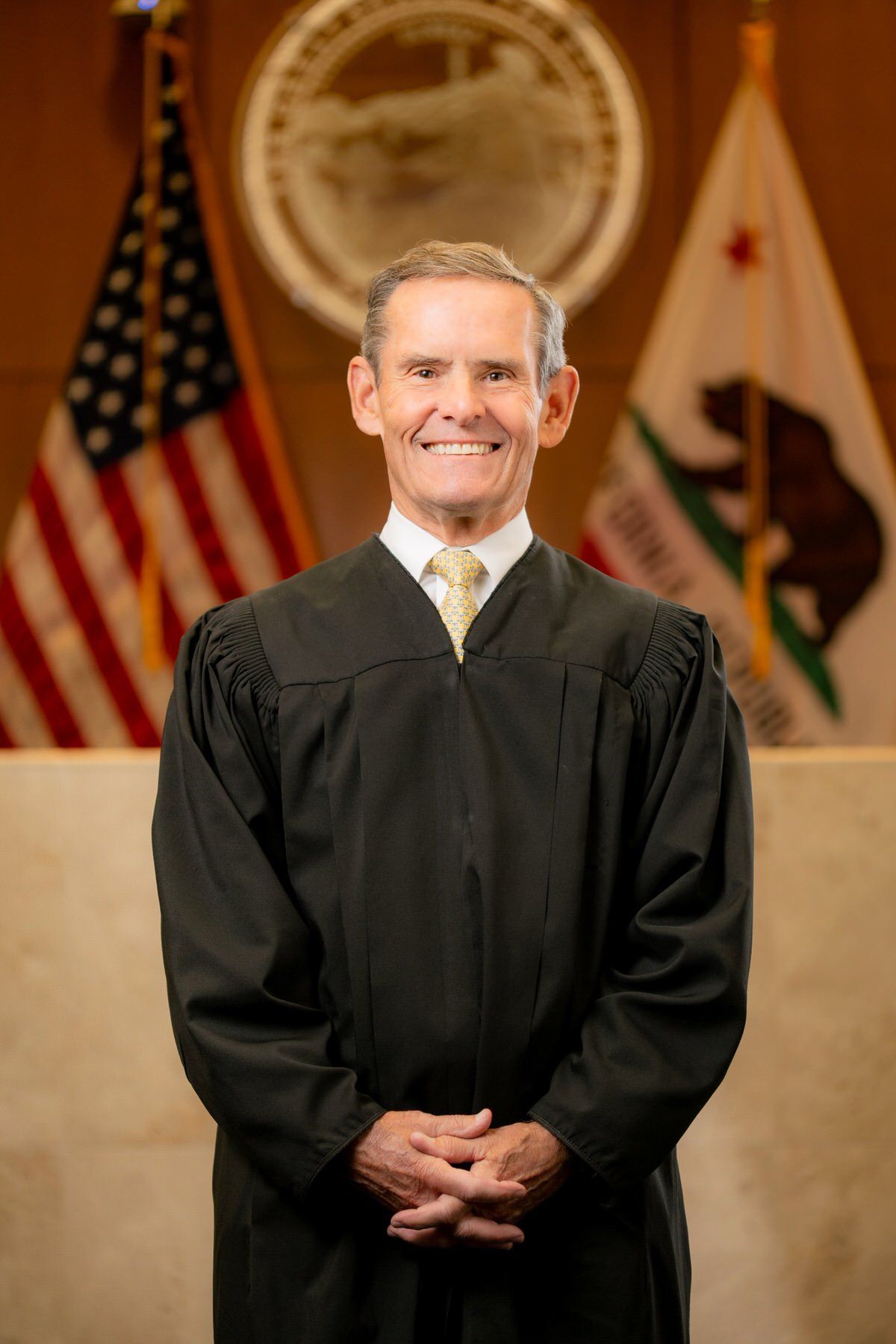 A man in a judge 's robe is standing in front of flags for a headshot.