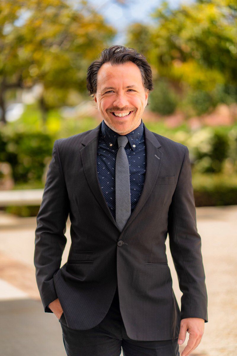 A man in a suit and tie is standing in a park with his hands in his pockets.