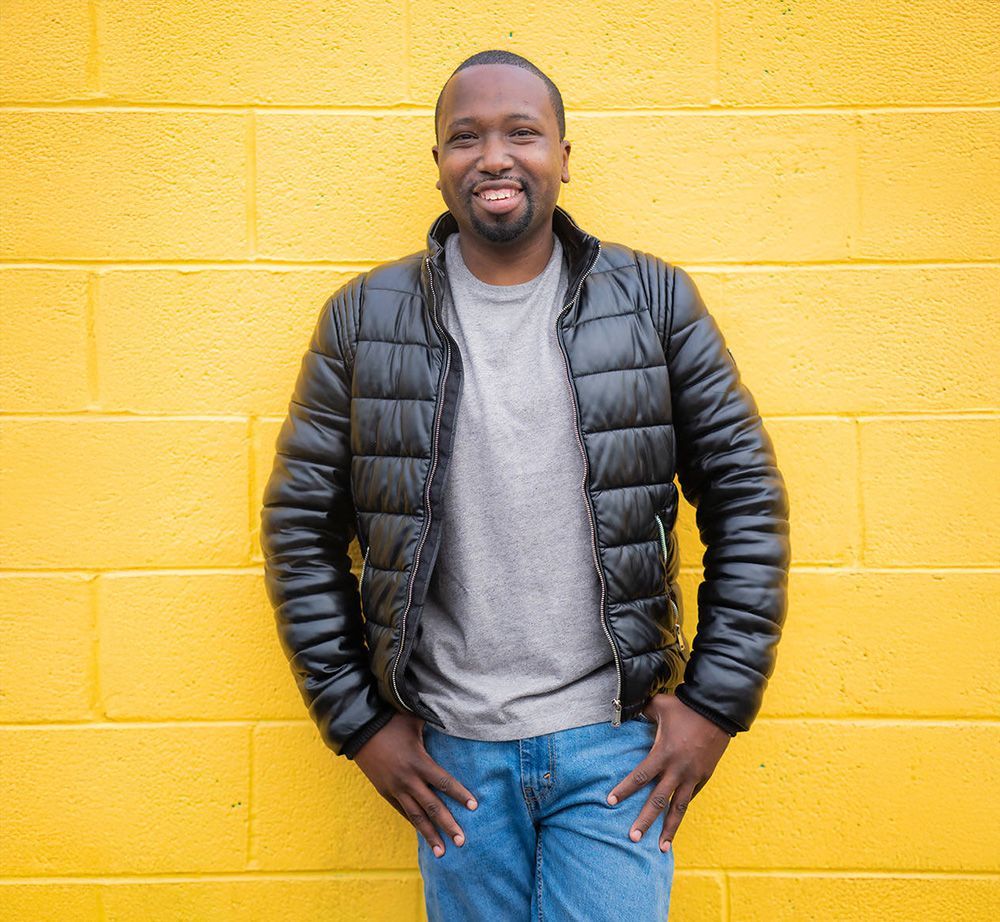 A man in a black jacket is standing in front of a yellow brick wall for a Coffee Meets Bagel App photo.