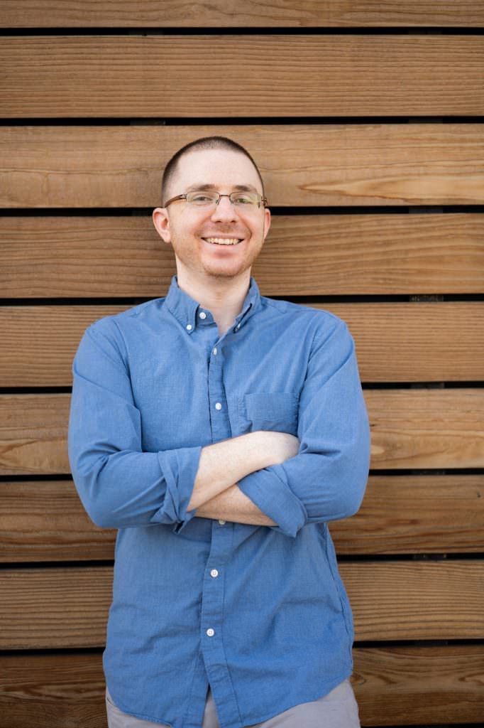 A man in a blue shirt and glasses is standing in front of a wooden wall with his arms crossed.