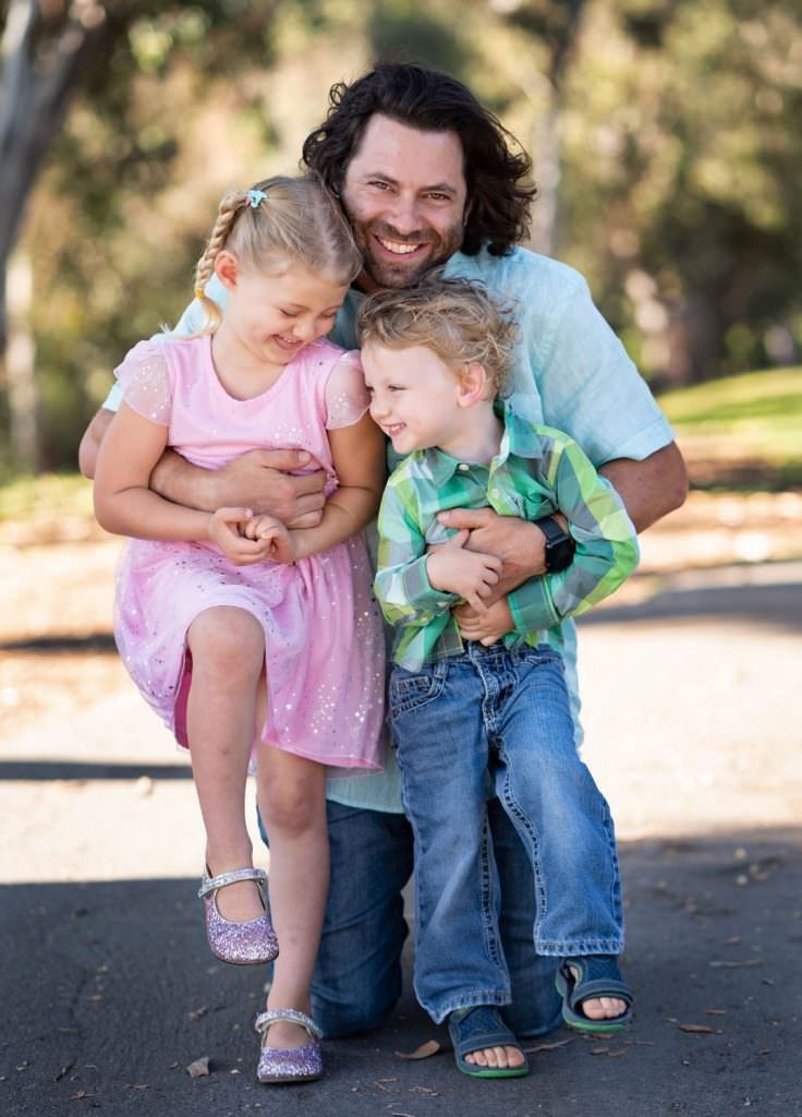 A man is kneeling down with two children in his arms in Huntington Central Park, Ca.