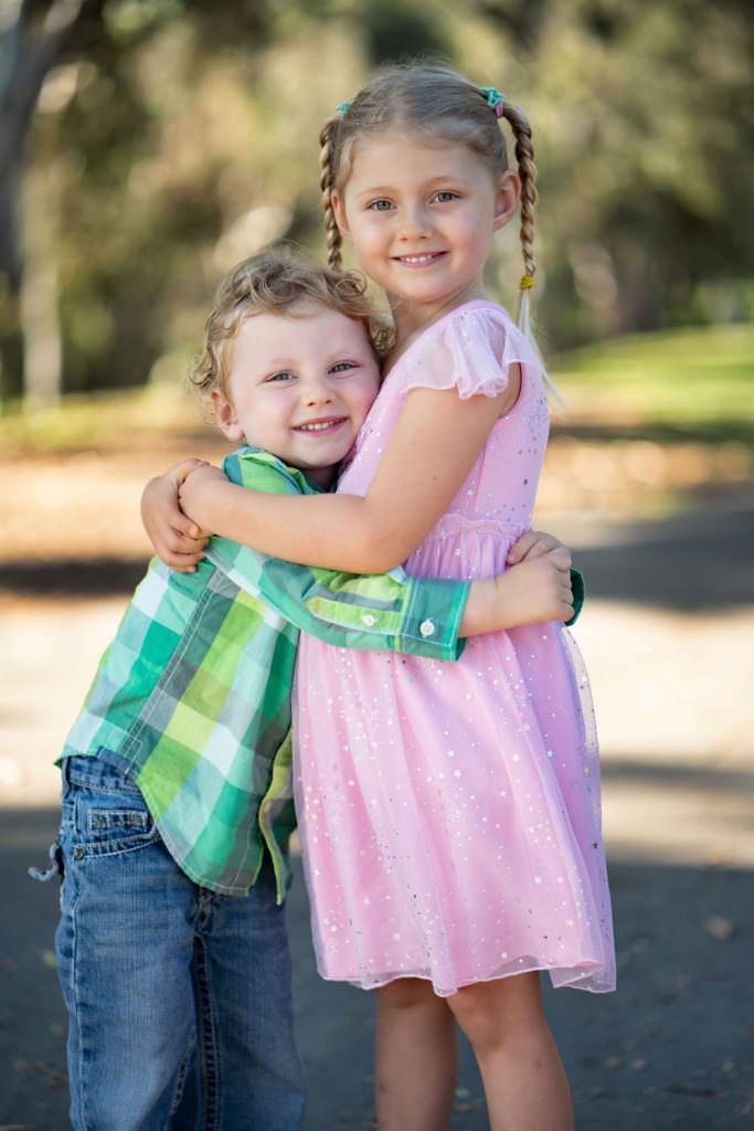 A young brother and sister are hugging each other in Huntington Beach park.