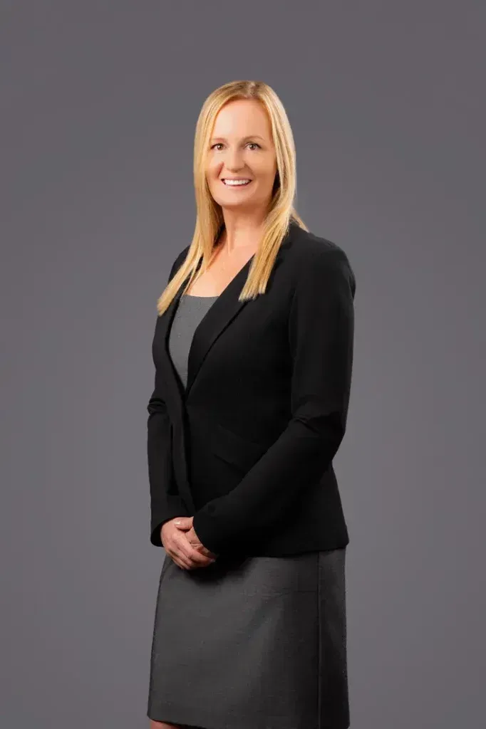 Female Corporate Headshot Example A woman in a black jacket and gray skirt is standing in front of a gray background.