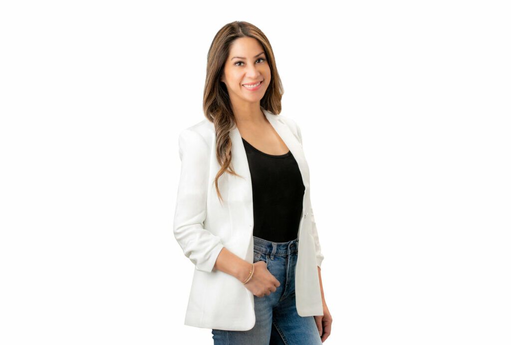 A woman wearing a white jacket and jeans is standing in front of a white background.