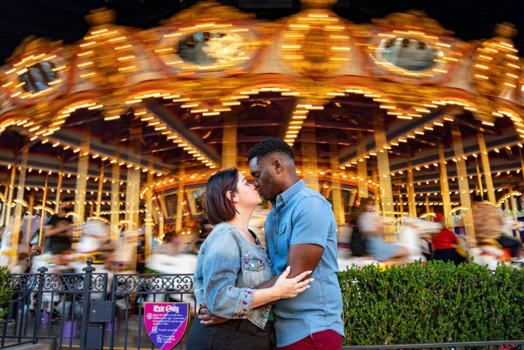 A man and a woman are kissing in front of a carousel.