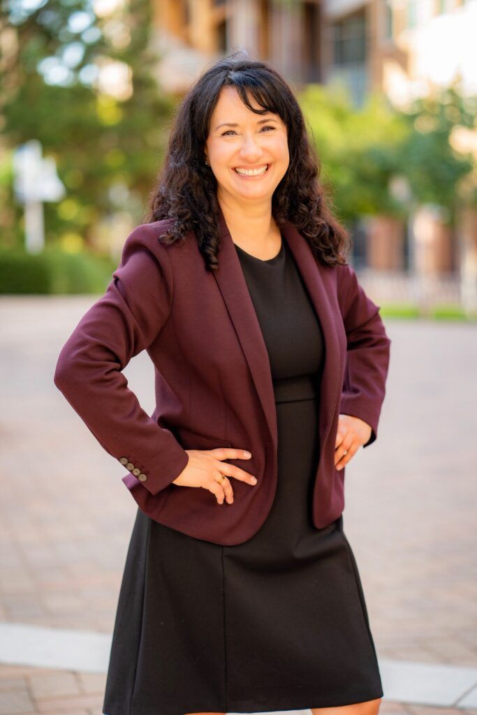 A woman in a black dress and a purple jacket is standing with her hands on her hips for a LinkedIn photo