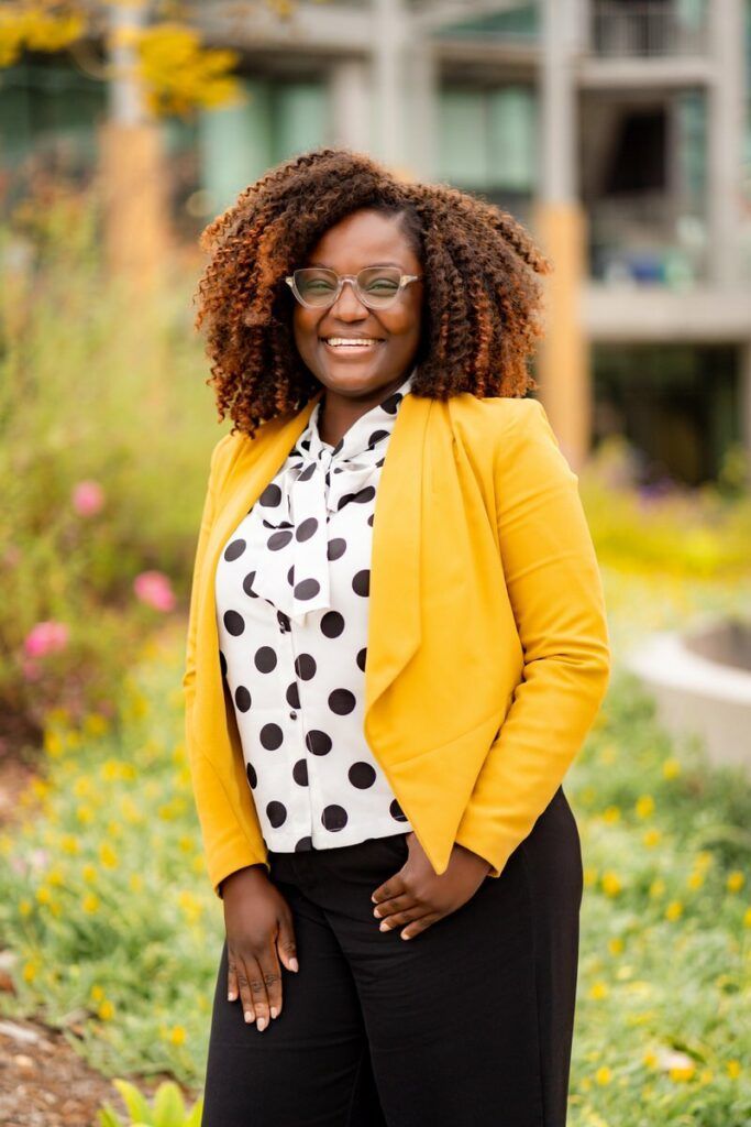 A woman wearing a yellow jacket and a polka dot shirt is standing in a garden.