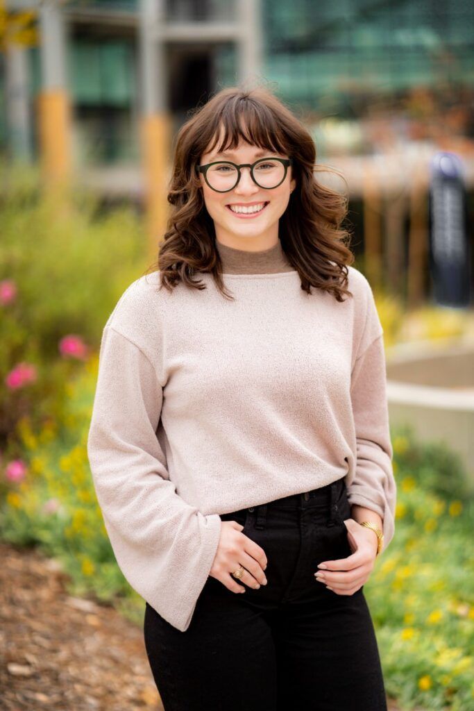 A woman wearing glasses and a sweater is standing in front of a flower bed.